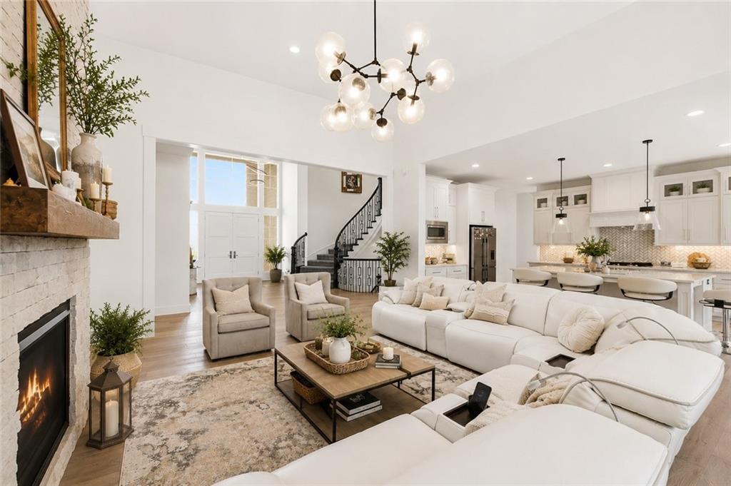 Open great room with a white sectional and two accent chairs facing a lit white-brick fireplace, a sputnik chandelier overhead, and the white kitchen and curved staircase beyond