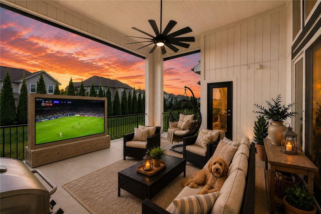 Covered patio at dusk with a black windmill ceiling fan, a large outdoor TV, wicker seating with striped cushions, a hanging egg chair, and a view of evergreen trees under a sunset sky