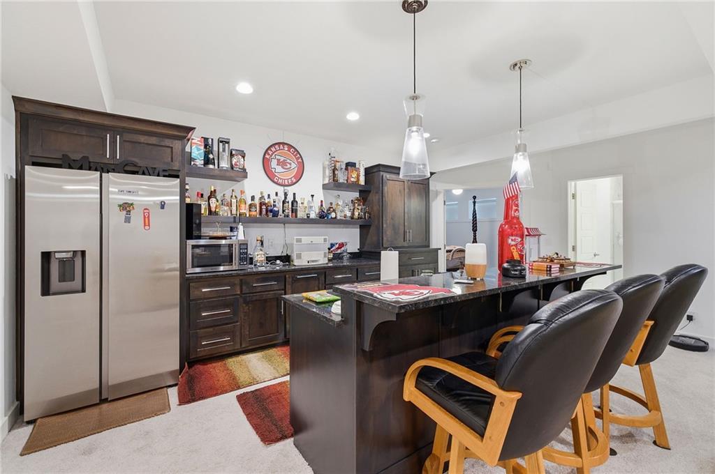 Lower-level wet bar with dark wood cabinetry, a full-size stainless refrigerator, open shelving of bottles, a dark granite bar with black leather swivel stools, and Kansas City Chiefs decor