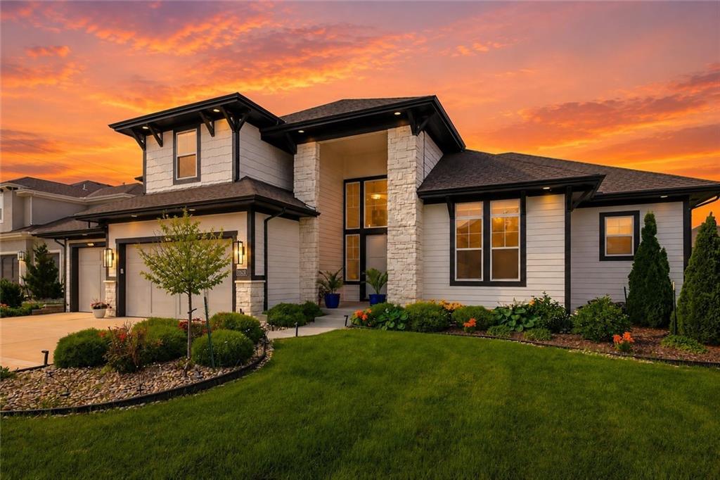 Twilight front elevation of 15620 Catalina St, a 1.5-story home with light siding, stone columns, and black trim, glowing at dusk under an orange sunset sky